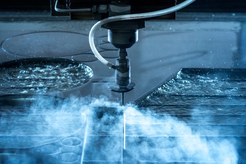 Waterjet cutting machine slicing through a metal sheet with a high-pressure stream of water and mist.